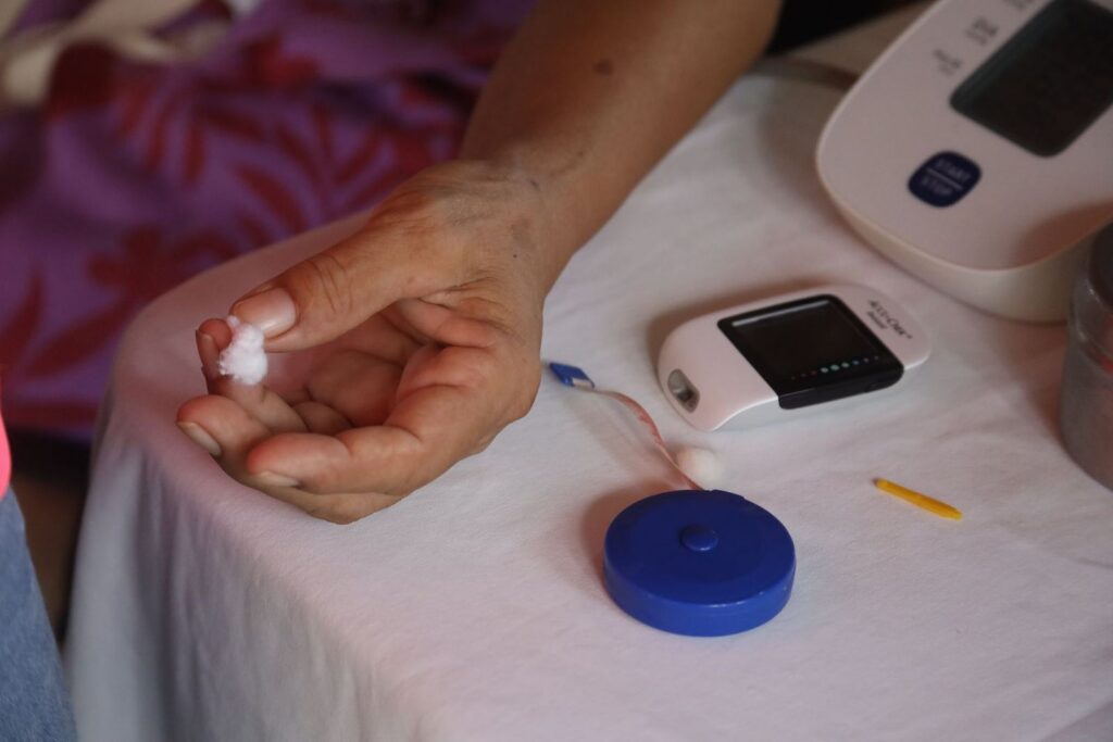 A hand holding a cotton ball beside a glucose meter and a blood pressure monitor on a table at home