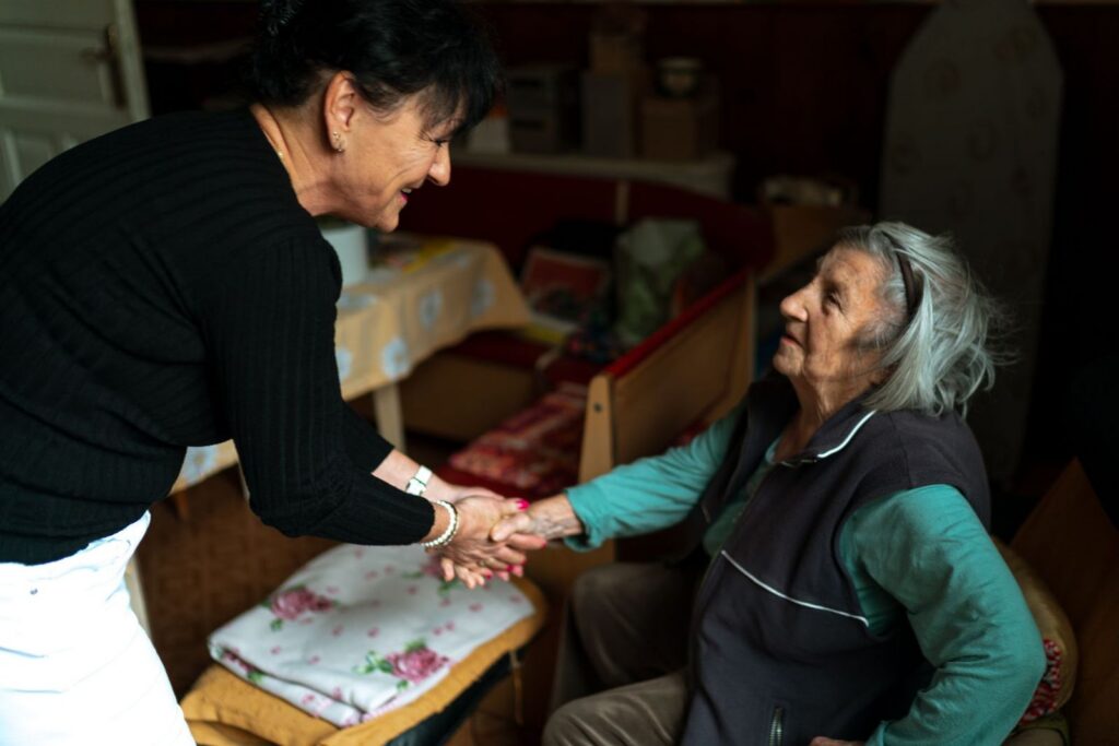 A woman leaning in to shake hands with a seated older woman in a warmly lit room at home