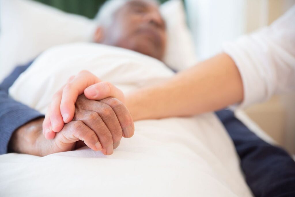 A carer gently holding the hand of an older person resting in bed