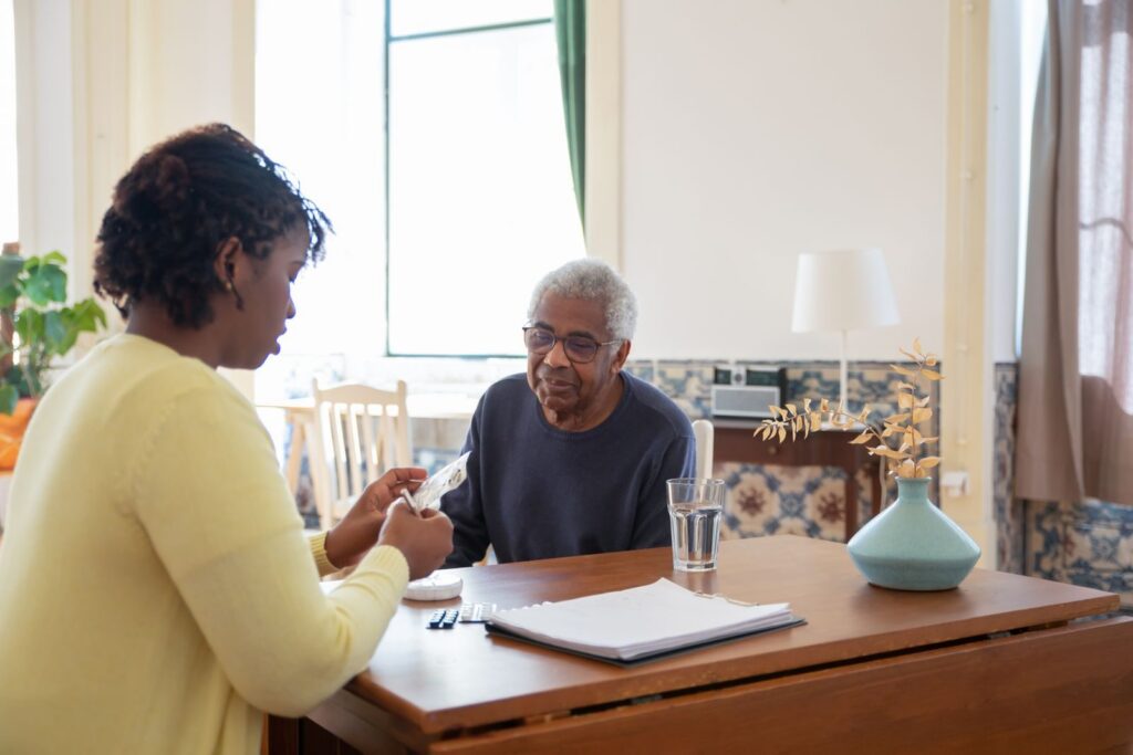 A carer helping an older man with his medication at a table in a sunlit living room