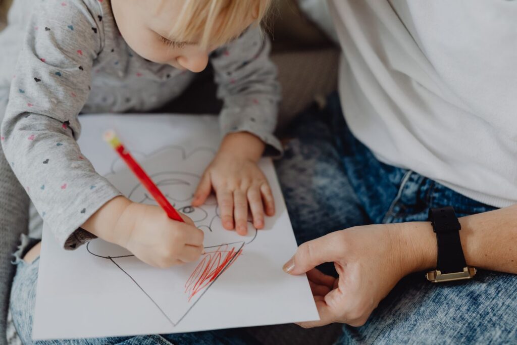 A young child colouring a drawing with a red pencil while an adult sits beside them