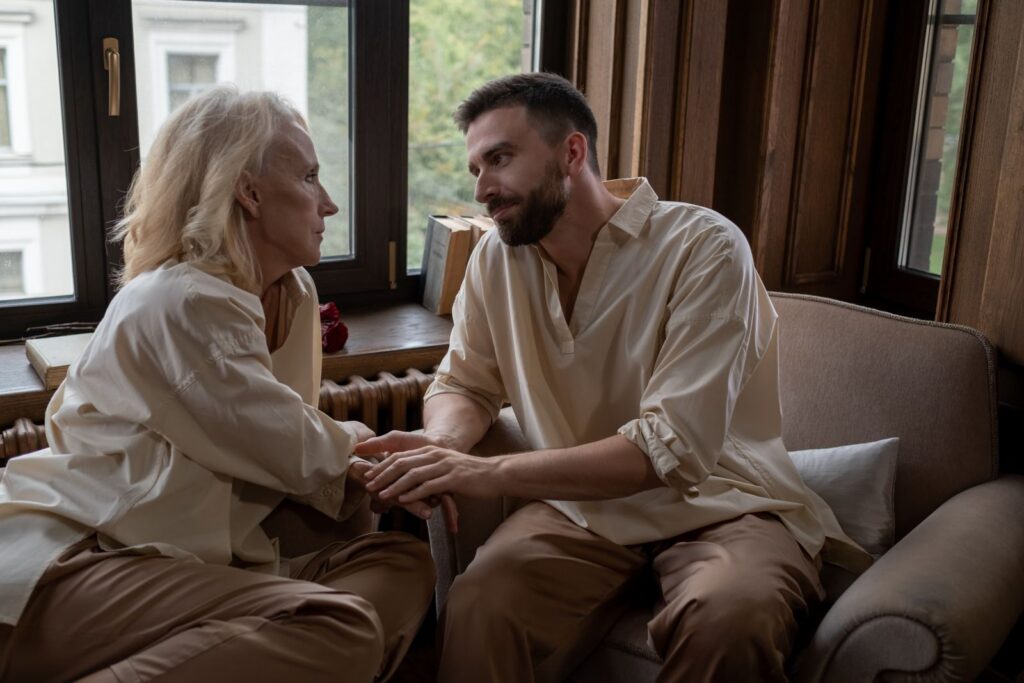 An older woman and a younger man holding hands and talking by a window at home