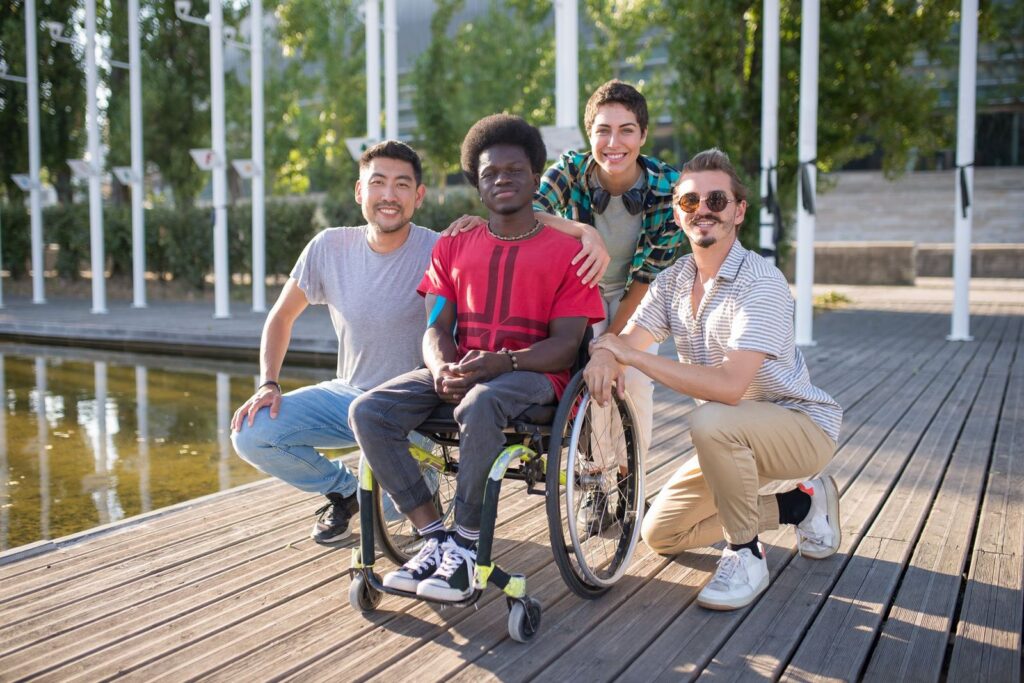 A group of four friends smiling for the camera on a timber deck beside a pond, one of them in a wheelchair