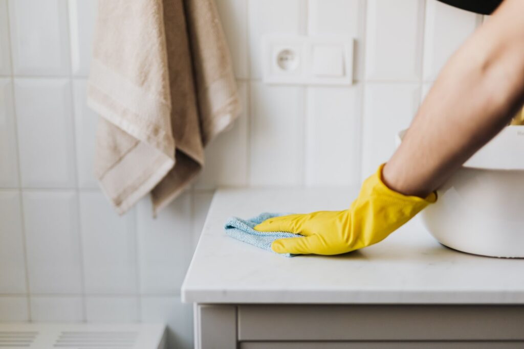 A hand in a yellow cleaning glove wiping a bathroom counter with a cloth