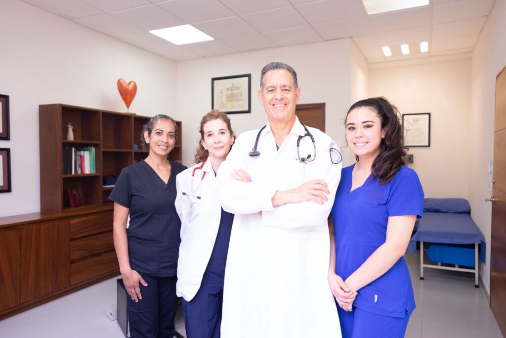 Five healthcare professionals in scrubs and white coats standing together and smiling in a clinic room