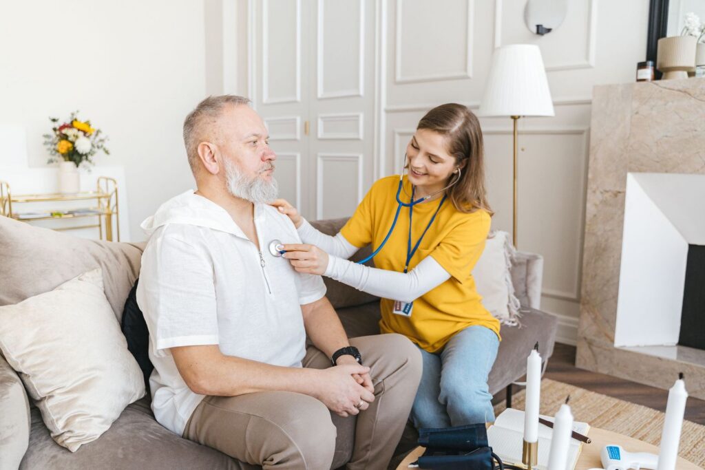 A healthcare worker in a yellow top listening to a man's chest with a stethoscope as they sit together on a sofa at home