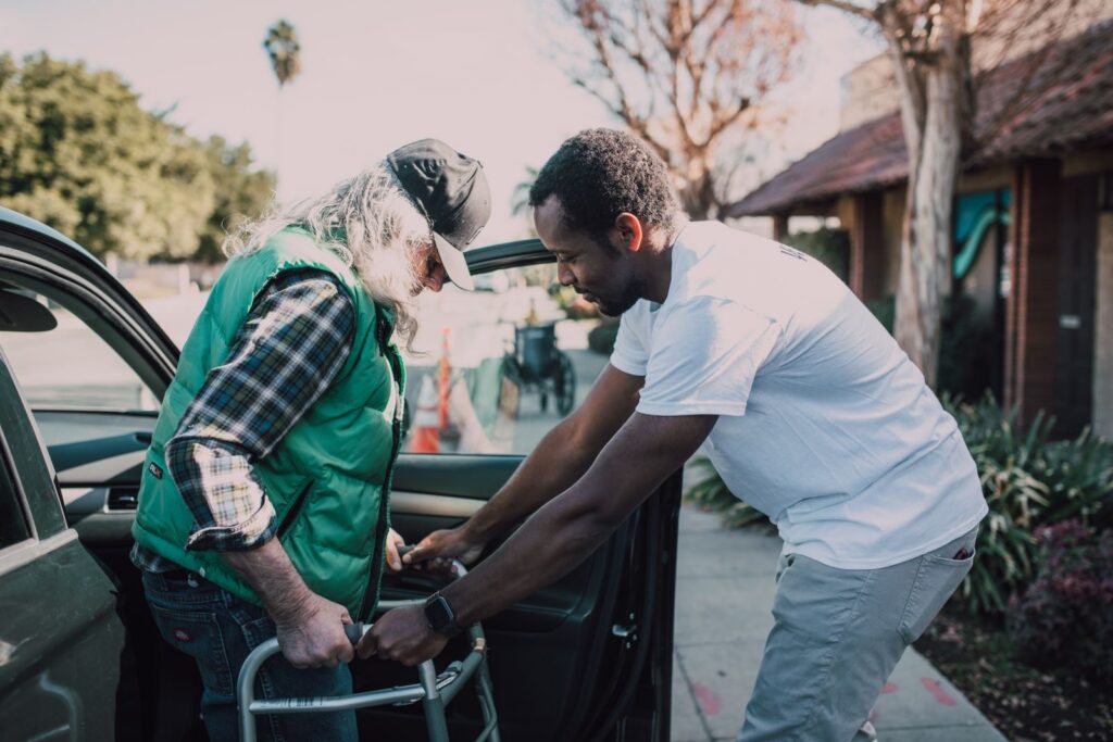 A man helping an older man with a walker get into a parked car