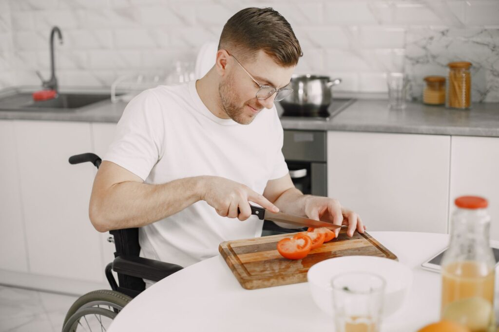 A man in a wheelchair slicing a tomato on a chopping board in a bright kitchen