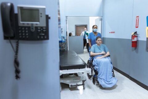 A nurse in scrubs and a mask pushing a patient in a wheelchair along a hospital corridor