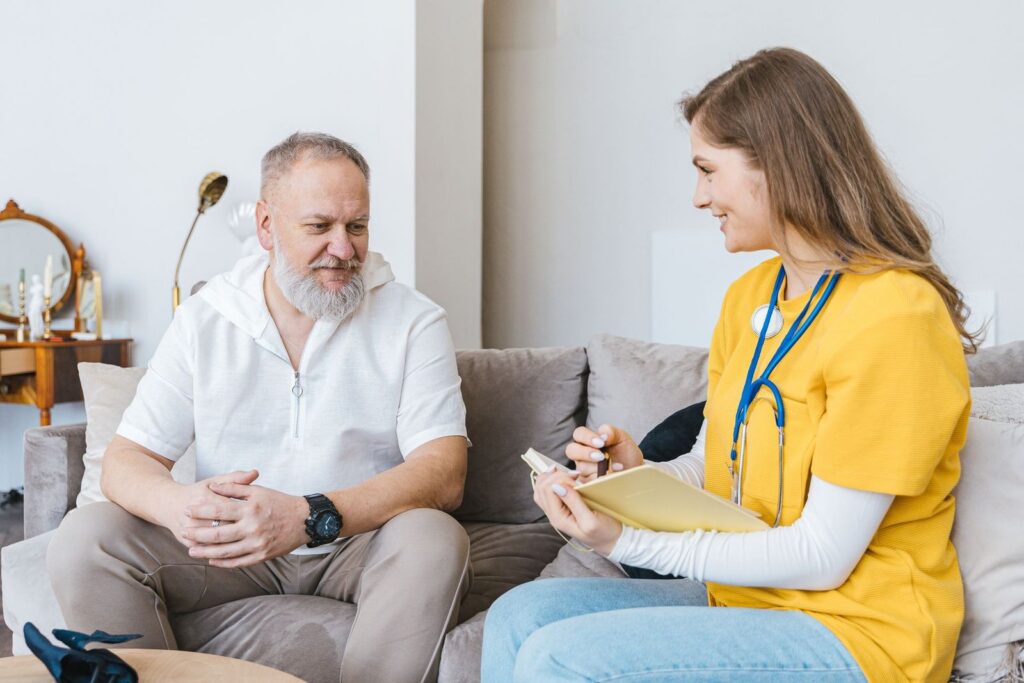 A woman in yellow scrubs writing on a clipboard while chatting with a smiling man on a sofa