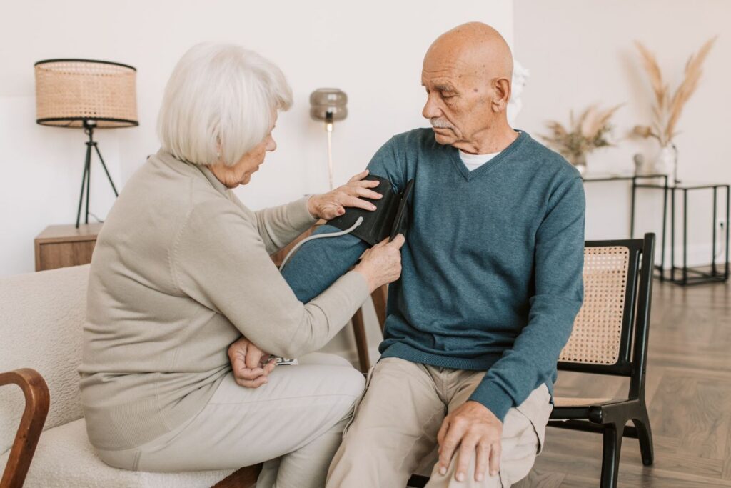 An older woman fitting a blood pressure cuff on an older man as they sit together in a bright living room at home