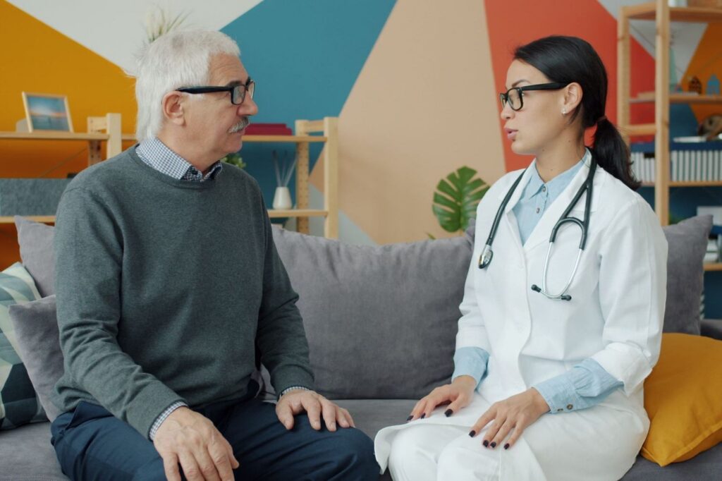 An older man in a green jumper talking with a woman in a white coat with a stethoscope, seated on a sofa