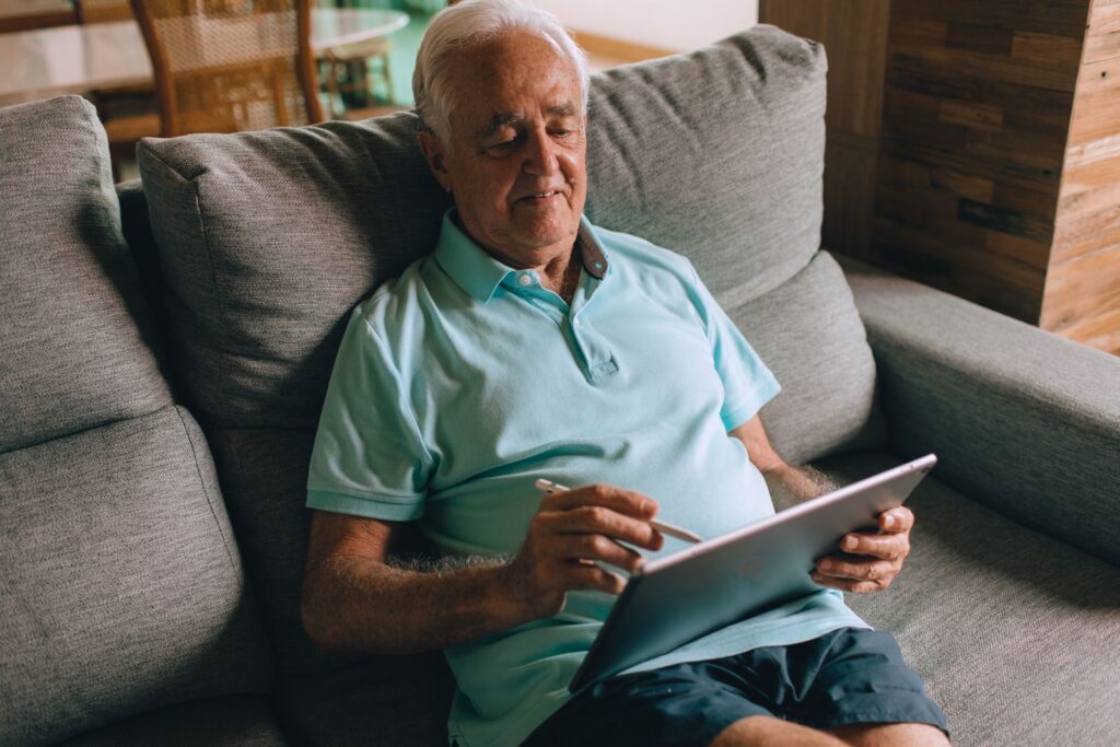 An older man smiling as he uses a tablet while sitting on a grey sofa at home