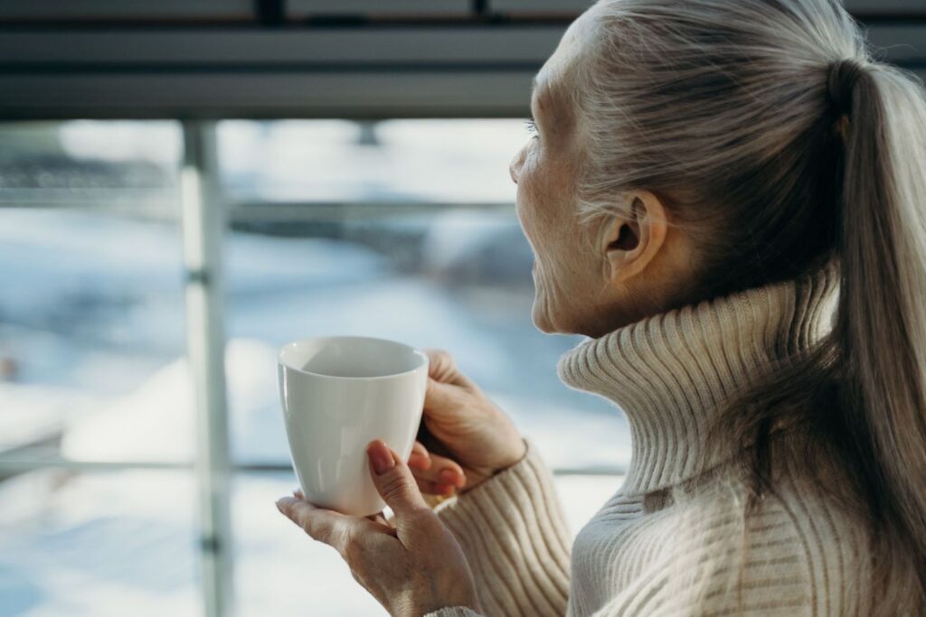 An older woman in a cream sweater holding a warm drink and looking out a window