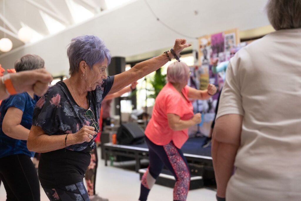 A group of older women dancing together in an indoor community space, one with her arm raised