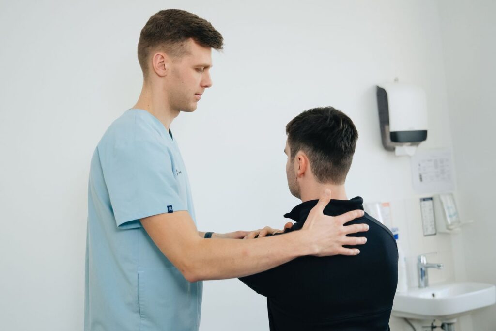 A clinician in light blue scrubs checking the shoulders of a patient in a bright treatment room