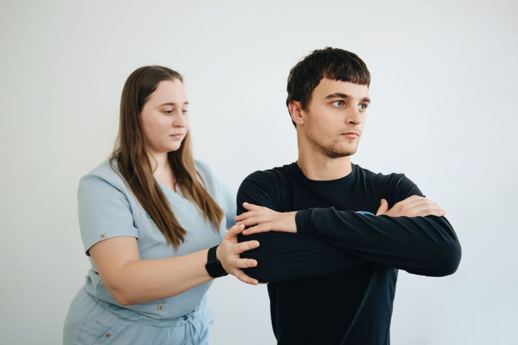 A clinician in light blue scrubs guiding a patient through an arm stretch in a bright clinic