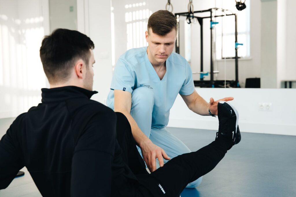 A physiotherapist in light blue scrubs guiding a patient through a leg stretch in a bright studio