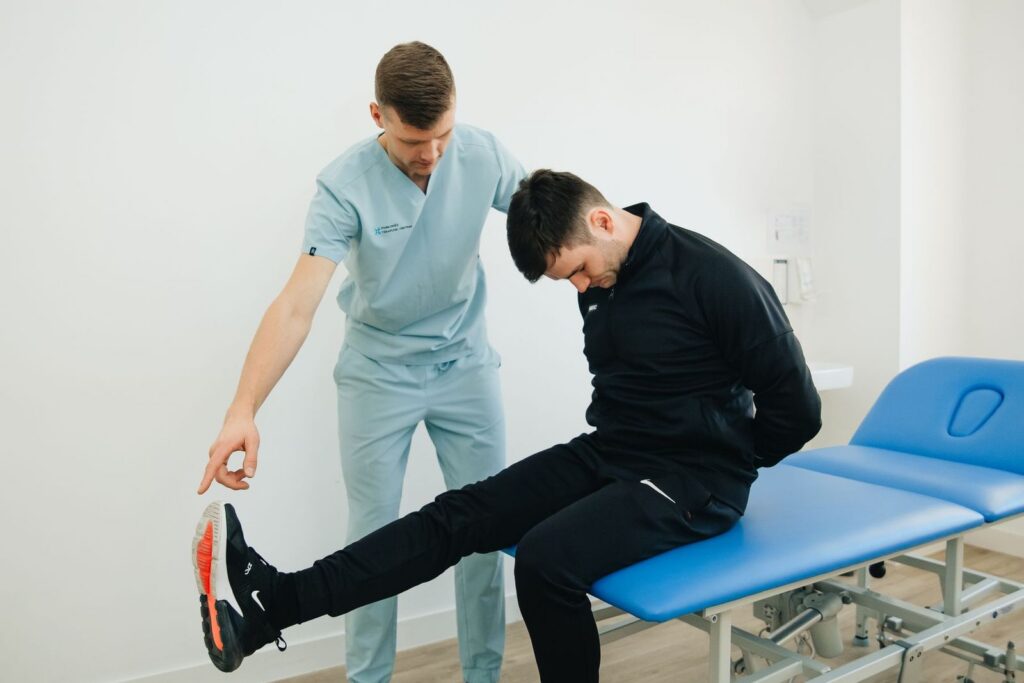A clinician in light blue scrubs guiding a patient through a seated leg stretch on a treatment table in a bright clinic