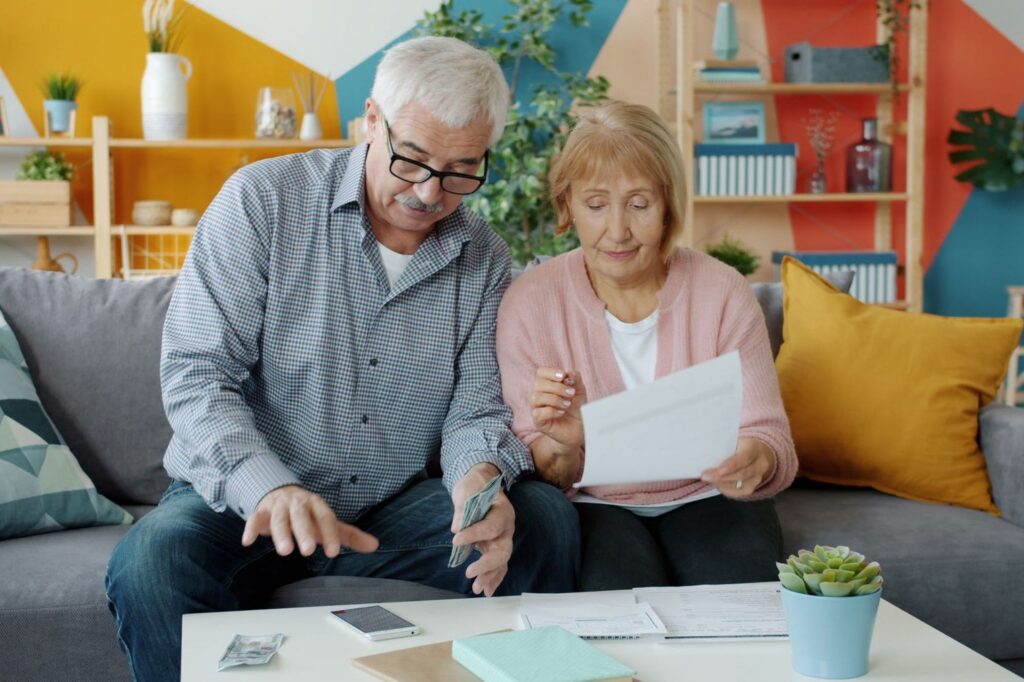 An older couple sitting on a sofa at home looking through paperwork together, with more documents on the coffee table