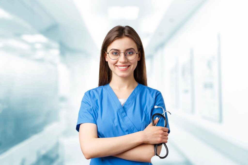 A smiling nurse in blue scrubs standing with her arms crossed, holding a stethoscope
