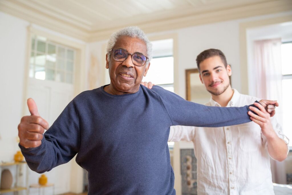A smiling older man giving a thumbs up while a younger man steadies his outstretched arm in a bright living room