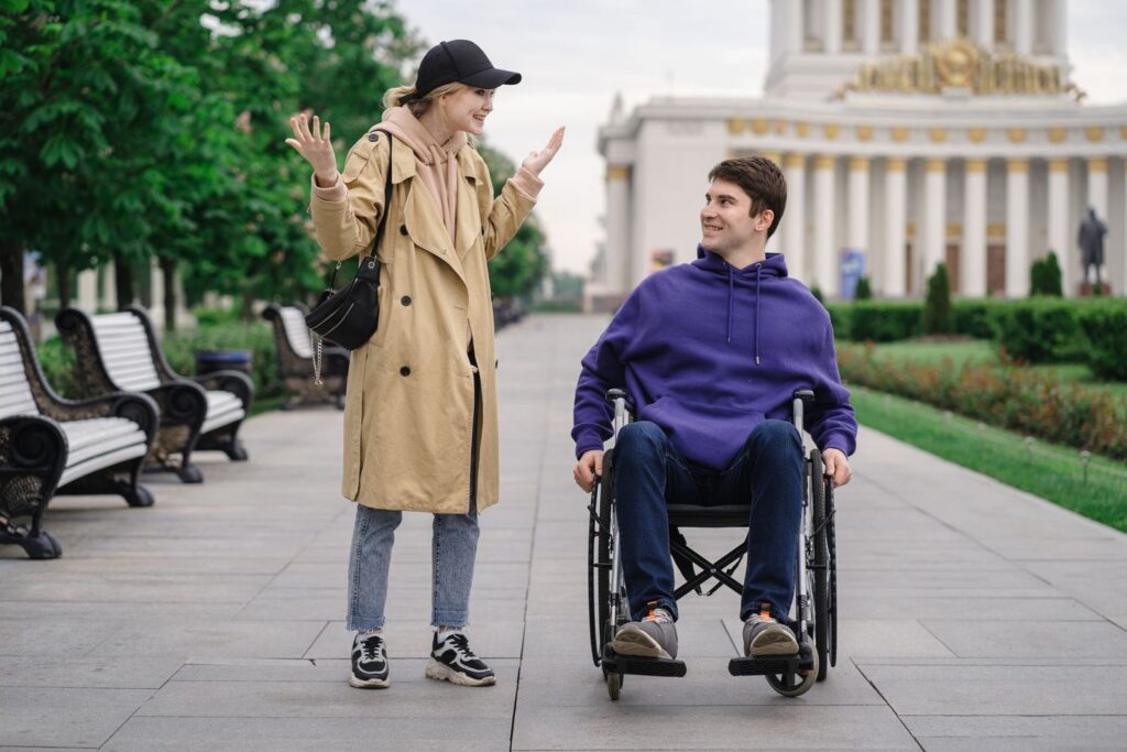 A woman and a man in a wheelchair talking and smiling together on a tree-lined path in a park
