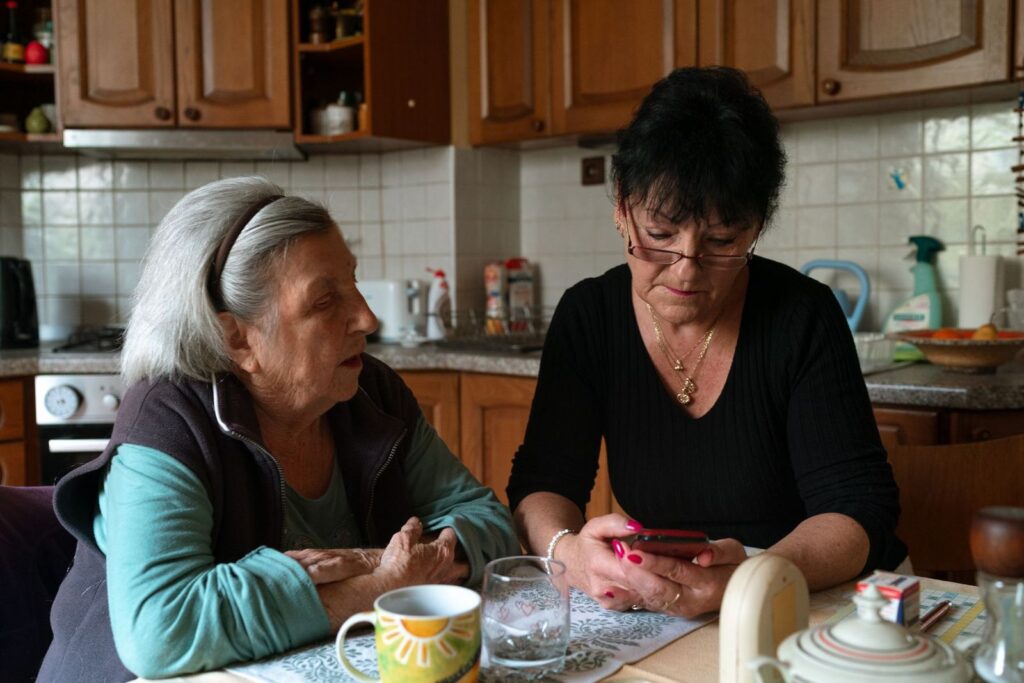 Two women sitting at a kitchen table talking while one of them uses a phone, with paperwork and mugs of tea in front of them