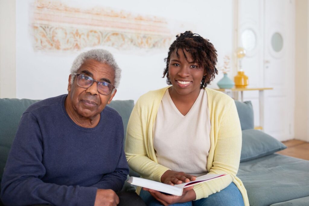 A smiling woman in a yellow cardigan holding an open book beside an older man on a sofa at home