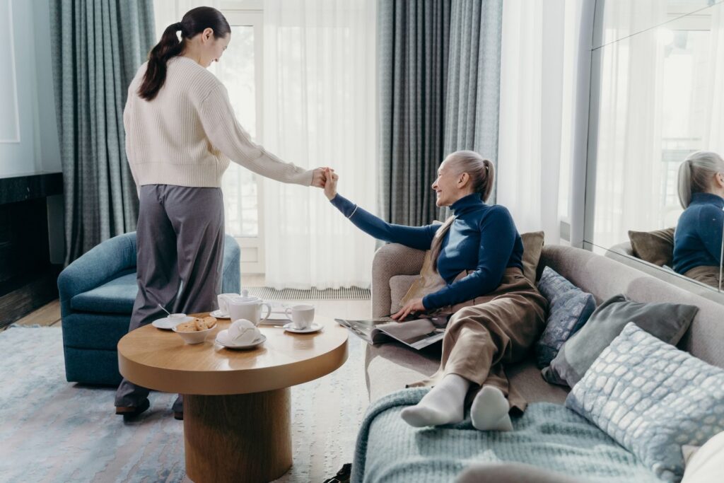 A woman holding the hand of an older woman seated on a sofa in a bright living room