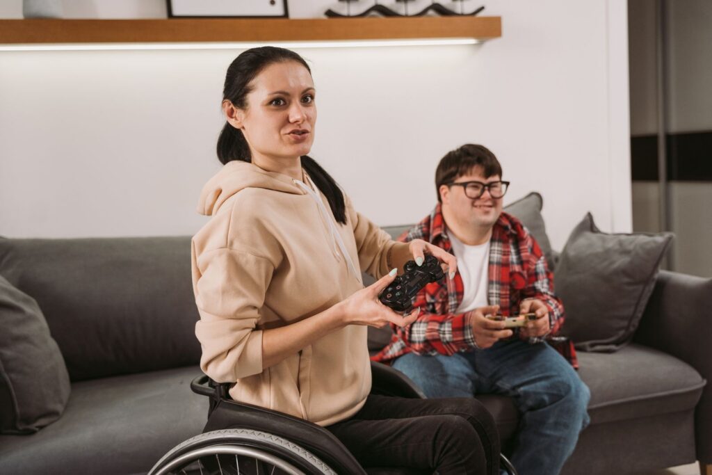 A woman in a wheelchair and a man on a sofa sitting together holding game controllers