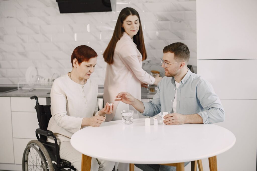 A woman in a wheelchair at a kitchen table being helped with her medication by two people