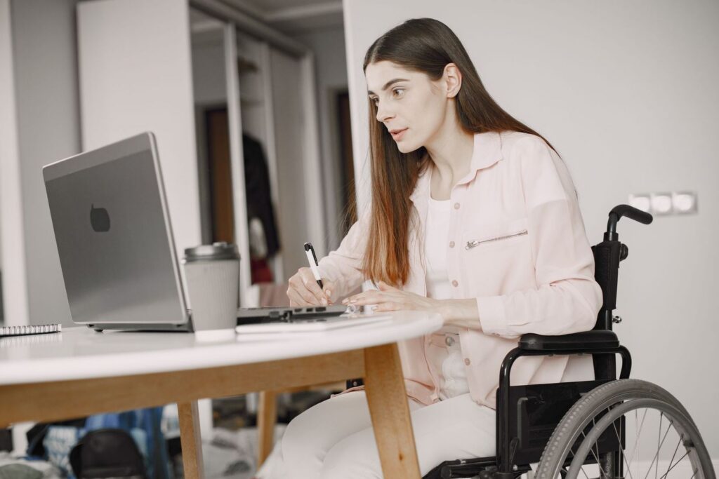 A woman in a wheelchair sitting at a round table, writing notes beside an open laptop