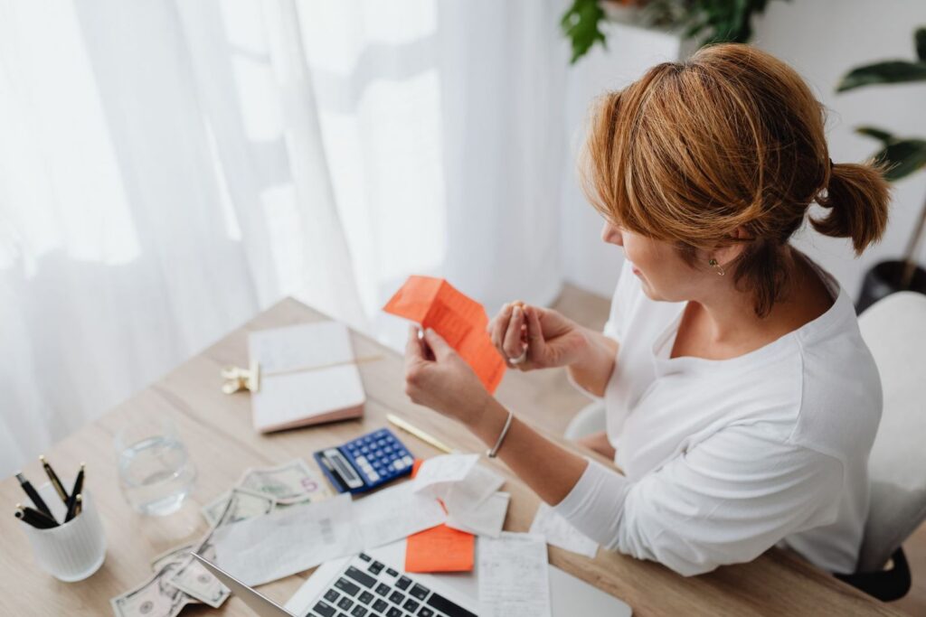 A woman at a home desk sorting through bills and receipts, with a calculator and laptop beside her