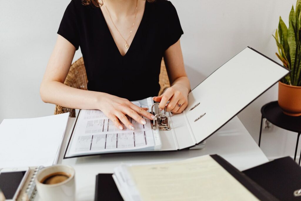 A woman at a desk filing forms into an open ring binder, with a cup of coffee beside her