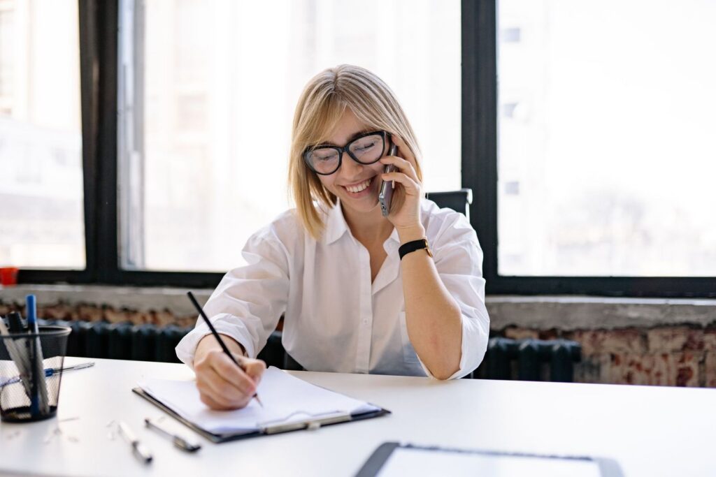 A smiling woman with glasses talking on the phone while writing notes on a clipboard at a desk