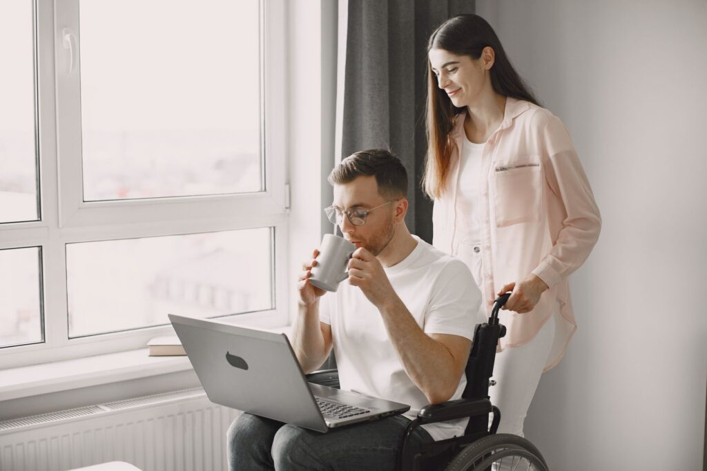 A woman standing beside a man in a wheelchair who is drinking from a cup with a laptop on his lap, by a bright window