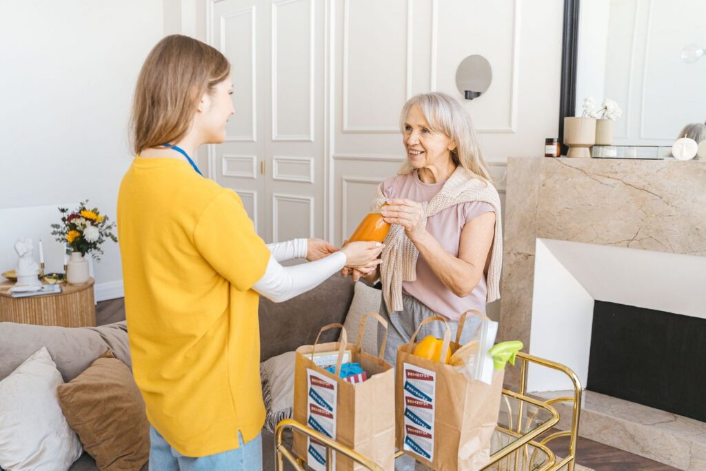 A young woman in a yellow top handing groceries to a smiling older woman in her living room