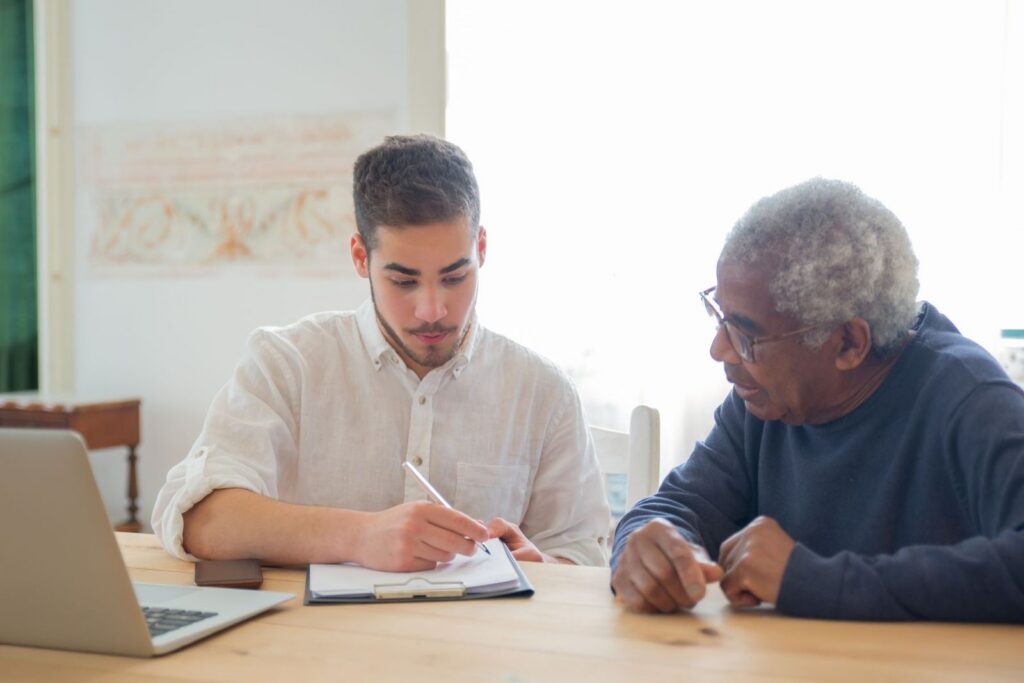 A young man writing on a clipboard while an older man sits beside him at a wooden table, going through documents together